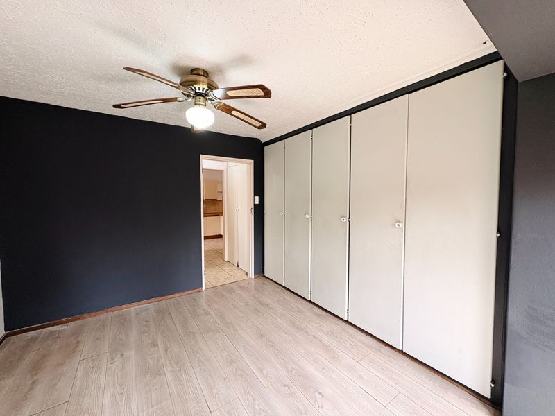 A spacious bedroom featuring light laminate flooring, a ceiling fan, and extensive built-in cupboard storage, leading through to an adjacent room.