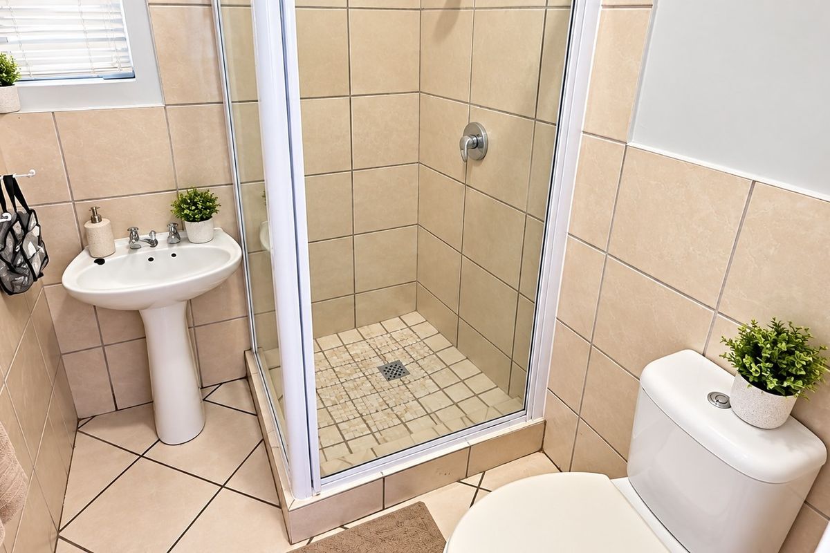 A compact, well-maintained bathroom featuring a pedestal basin, a glass-enclosed shower, and a standard toilet with neutral wall and floor tiling.