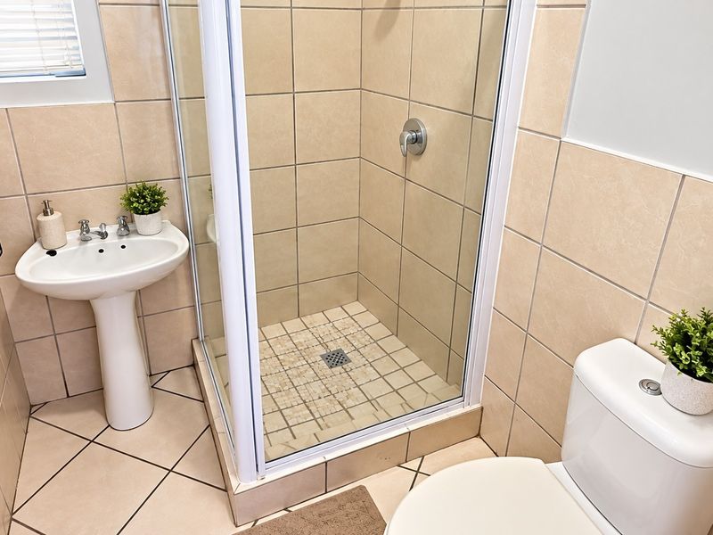 A compact, well-maintained bathroom featuring a pedestal basin, a glass-enclosed shower, and a standard toilet with neutral wall and floor tiling.