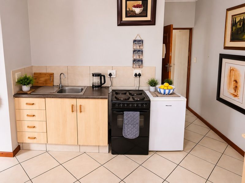 A compact kitchen area featuring light wood-finish cabinetry, a stainless steel sink, tiled backsplash, and a freestanding electric stove with ceramic floor tiles.