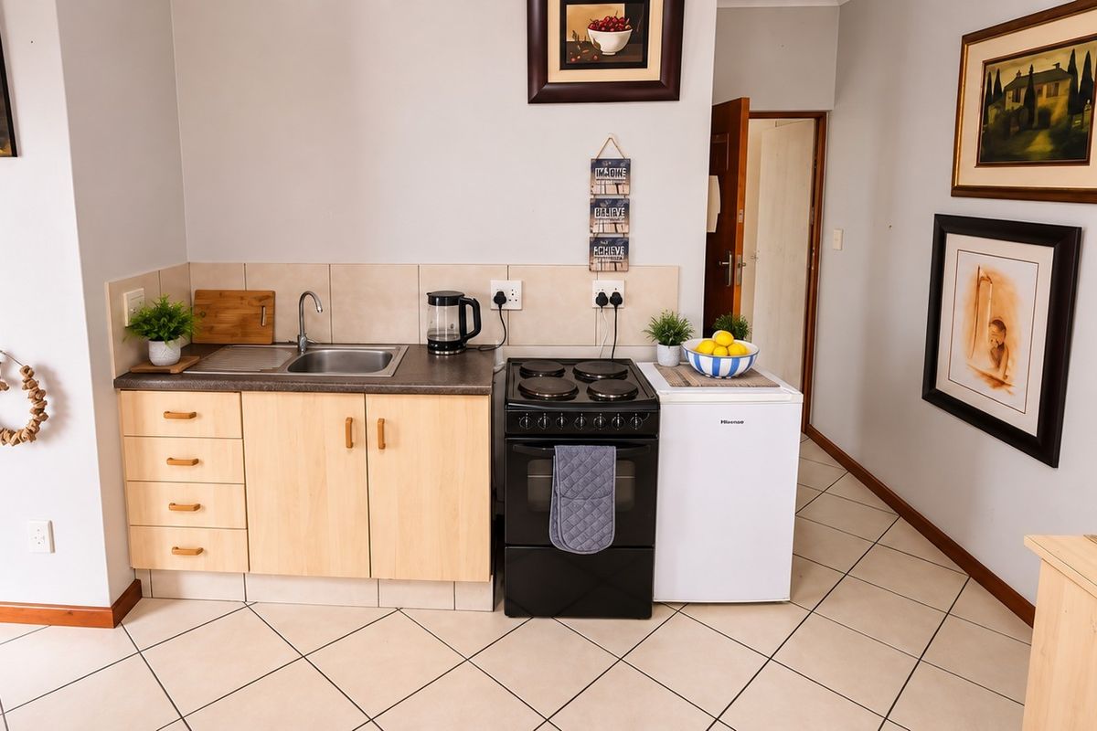 A compact kitchen area featuring light wood-finish cabinetry, a stainless steel sink, tiled backsplash, and a freestanding electric stove with ceramic floor tiles.