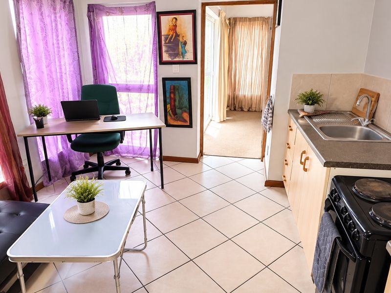 Open-plan living area featuring a compact kitchen with built-in cabinetry, electric stove, and tiled flooring, seamlessly connected to a versatile living and workspace.