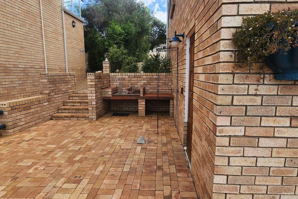 A paved rear courtyard featuring a built-in brick braai area, retaining walls, and steps leading to an elevated garden section.