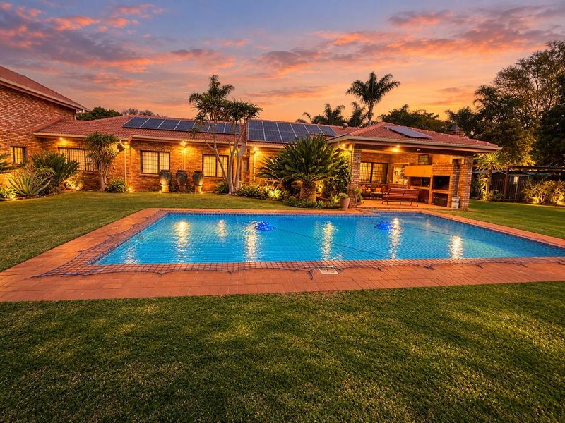 A beautifully landscaped backyard featuring a swimming pool, covered patio with built-in braai, and solar panels on the tiled roof at sunset.