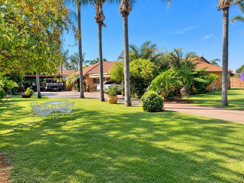 Spacious front garden with a well-maintained lawn, mature palm trees, brick driveway, and a view of the main house facade and carport.