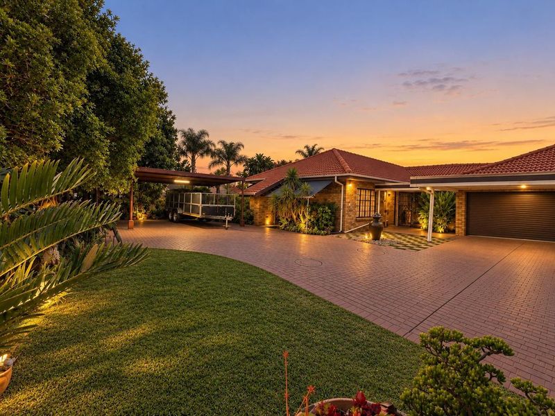 A well-maintained brick residence featuring a spacious paved driveway, double garage, carport, and beautifully landscaped front garden at sunset.