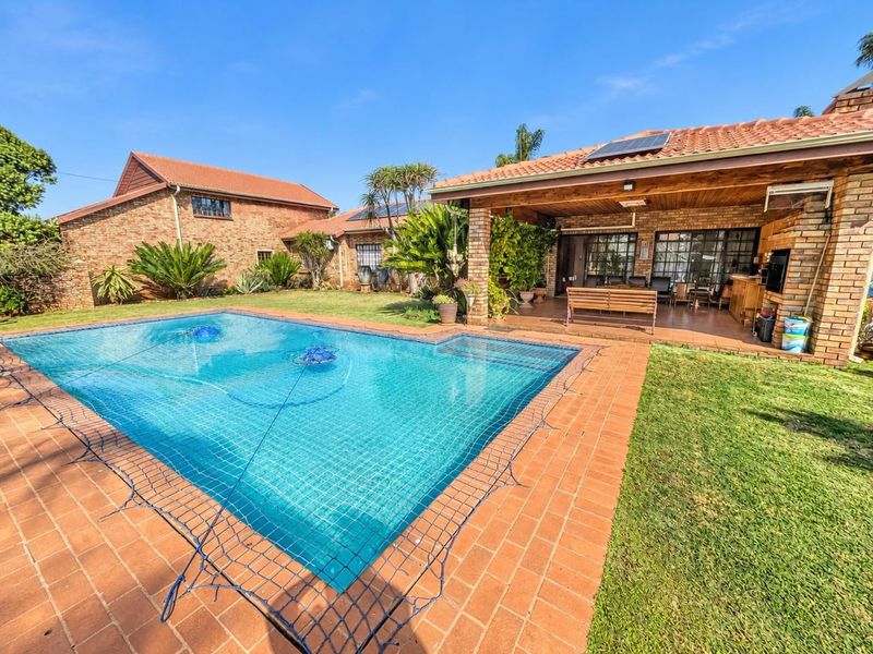 A well-maintained outdoor area featuring a swimming pool with a safety net, adjacent to a covered brick patio and landscaped garden.