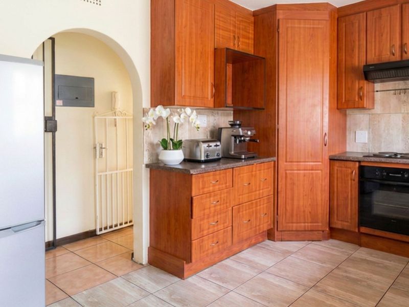A functional kitchen featuring warm-toned wooden cabinetry, built-in oven and hob, tiled backsplash, and durable floor tiles, leading through an arched doorway.