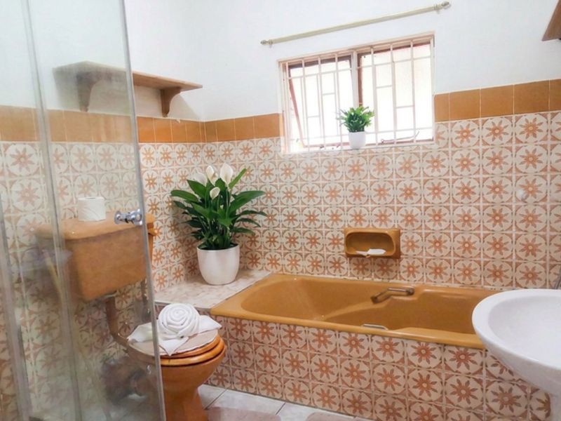 A functional bathroom featuring a bathtub, pedestal basin, toilet, and shower enclosure with vintage-style patterned wall tiles and security bars on the window.