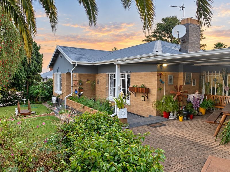 A well-maintained rear exterior view of a brick house featuring a paved patio area, carport, and established garden under a sunset sky.