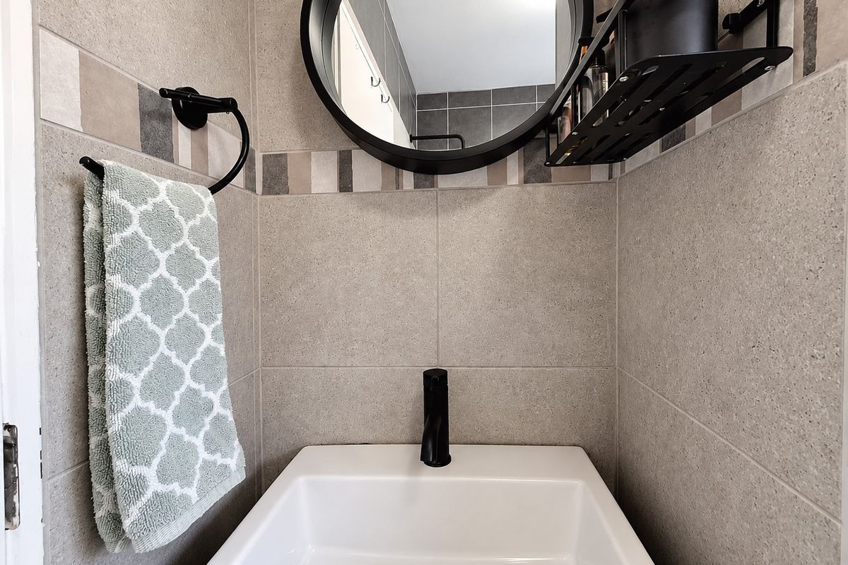 A modern bathroom vanity area featuring a white basin, black mixer tap, round mirror, and neutral-toned wall tiles with a decorative accent border.