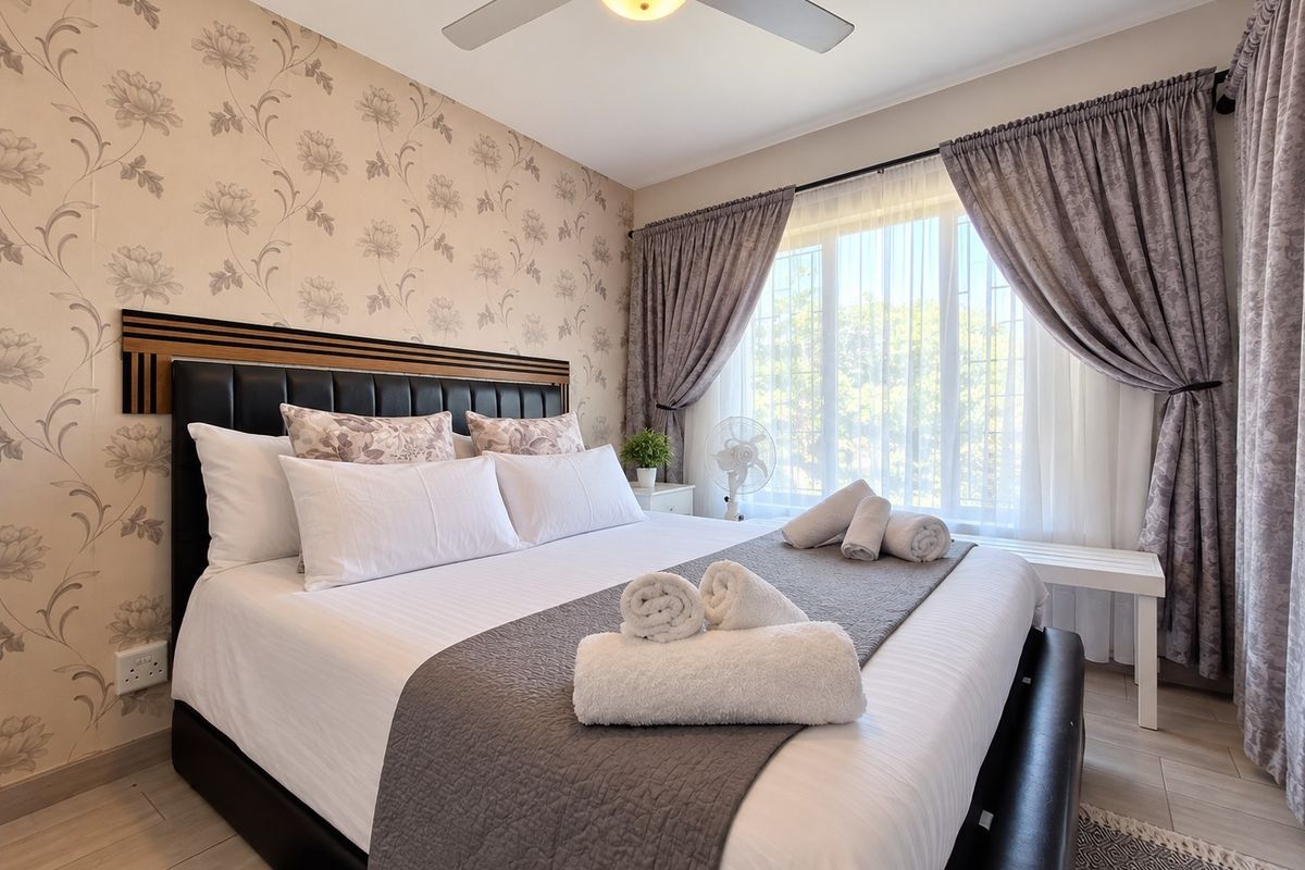 A well-maintained bedroom featuring patterned wallpaper, tiled flooring, and a ceiling fan, with large windows dressed in layered curtains.