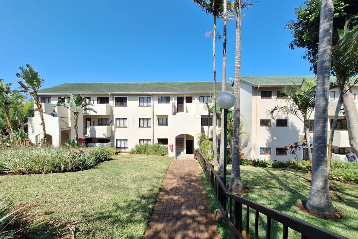 A view of the apartment complex exterior featuring a paved walkway leading to the building entrance, surrounded by a well-maintained garden and palm trees.