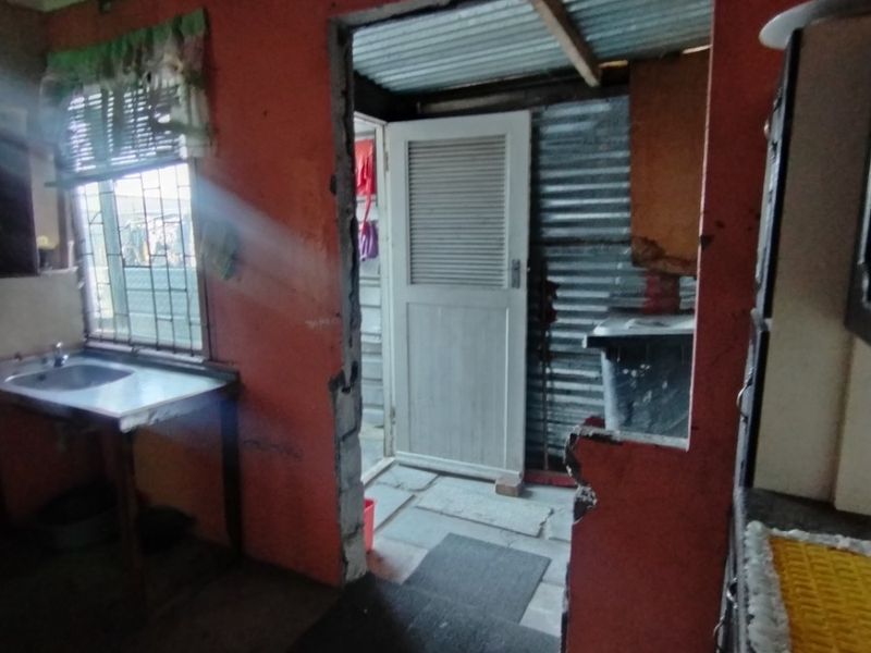 A dilapidated kitchen area featuring a stainless steel sink, significant ceiling water damage, and an open doorway leading to an exterior corrugated metal structure.