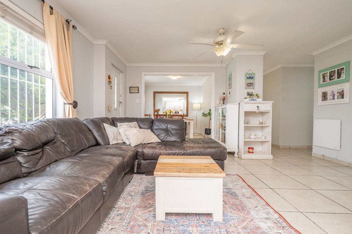 A comfortable living room featuring tiled flooring, a ceiling fan, and large windows with security bars, leading into an adjacent dining area.
