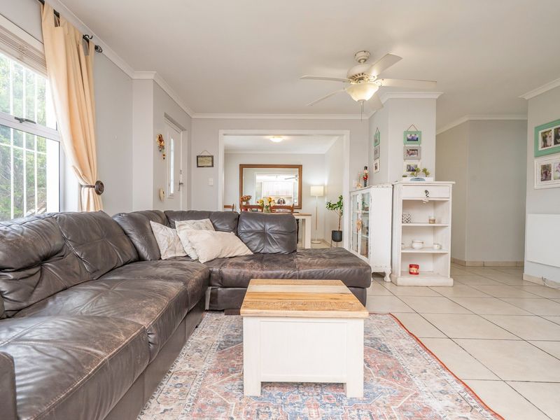 A comfortable living room featuring tiled flooring, a ceiling fan, and large windows with security bars, leading into an adjacent dining area.