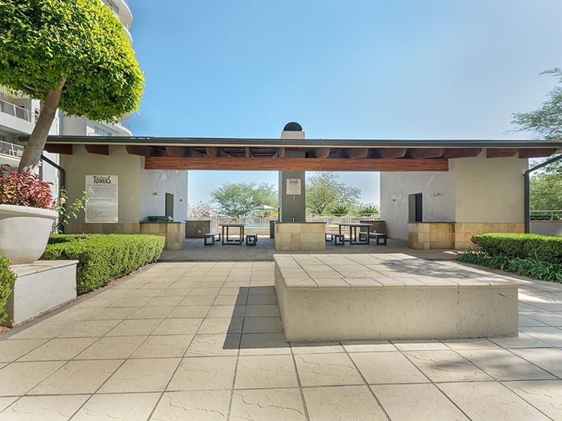 A well-maintained outdoor pool area featuring a covered patio with a built-in braai, paved terrace, and landscaped garden surroundings.