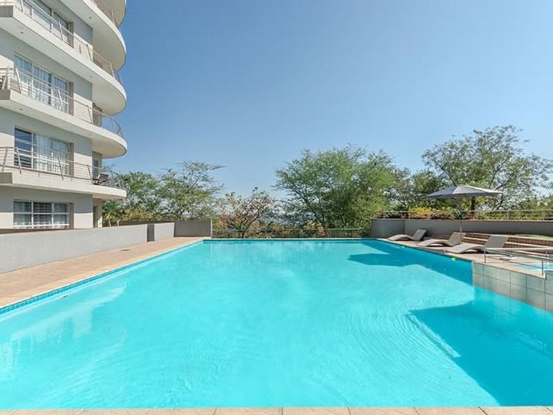 A well-maintained outdoor swimming pool area featuring a clear blue pool, paved surround, and views of the surrounding apartment building and greenery.