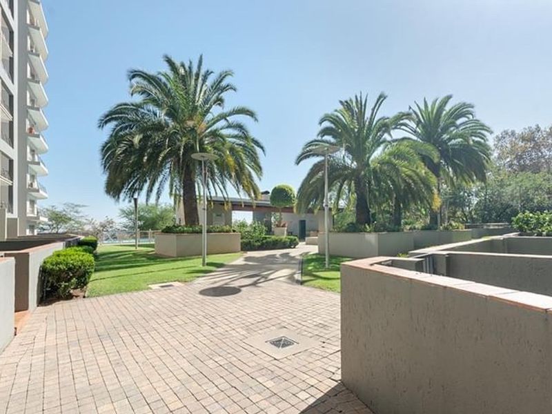 A well-maintained communal garden area featuring paved walkways, established palm trees, and structured retaining walls within the apartment complex grounds.