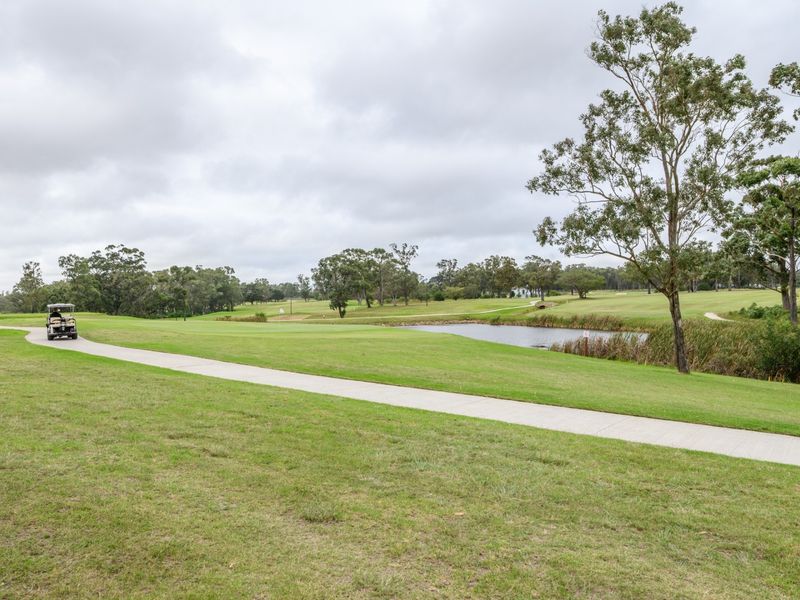 A scenic view of a well-maintained golf course featuring a concrete pathway, a small pond, and mature trees under an overcast sky.
