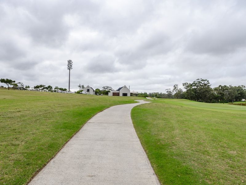 A view of a concrete pathway winding through a well-maintained grassy landscape with distant buildings and a telecommunications tower under a cloudy sky.