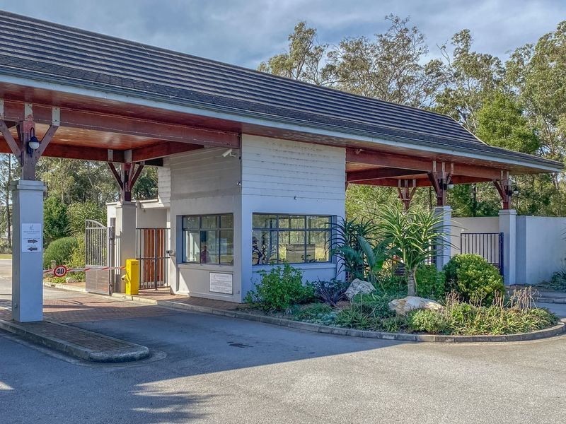 A well-maintained security gatehouse entrance featuring a boom gate, timber roof structure, and landscaped garden area at the estate access point.