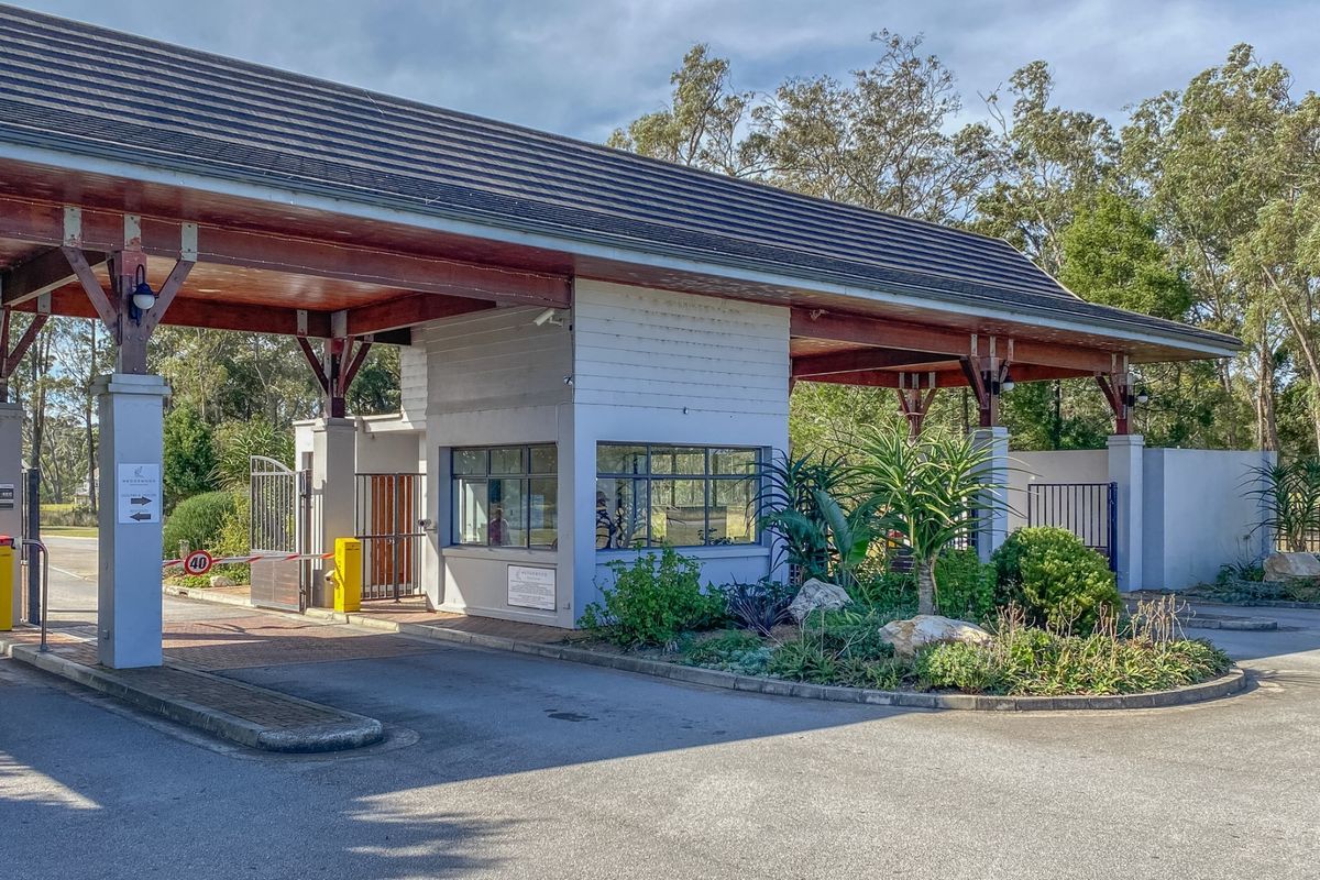 A well-maintained security gatehouse entrance featuring a boom gate, timber roof structure, and landscaped garden area at the estate access point.