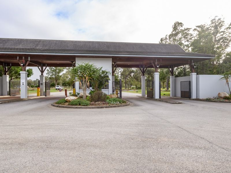 A well-maintained residential estate entrance featuring a covered security gatehouse, asphalt driveway, and landscaped traffic island.