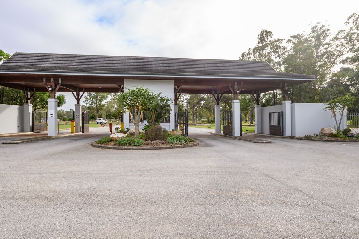 A well-maintained residential estate entrance featuring a covered security gatehouse, asphalt driveway, and landscaped traffic island.