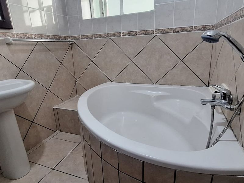 A functional bathroom featuring a corner bathtub with a handheld shower mixer, a pedestal basin, and neutral ceramic wall and floor tiling.