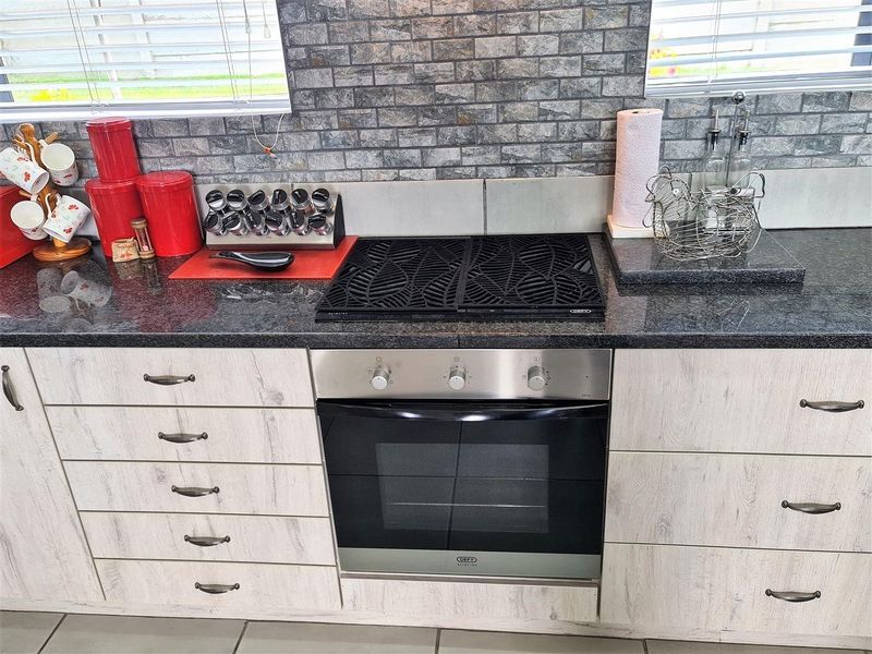 Modern kitchen featuring light wood-grain cabinetry, dark granite countertops, a built-in oven and hob, and a decorative stone-look tile backsplash.