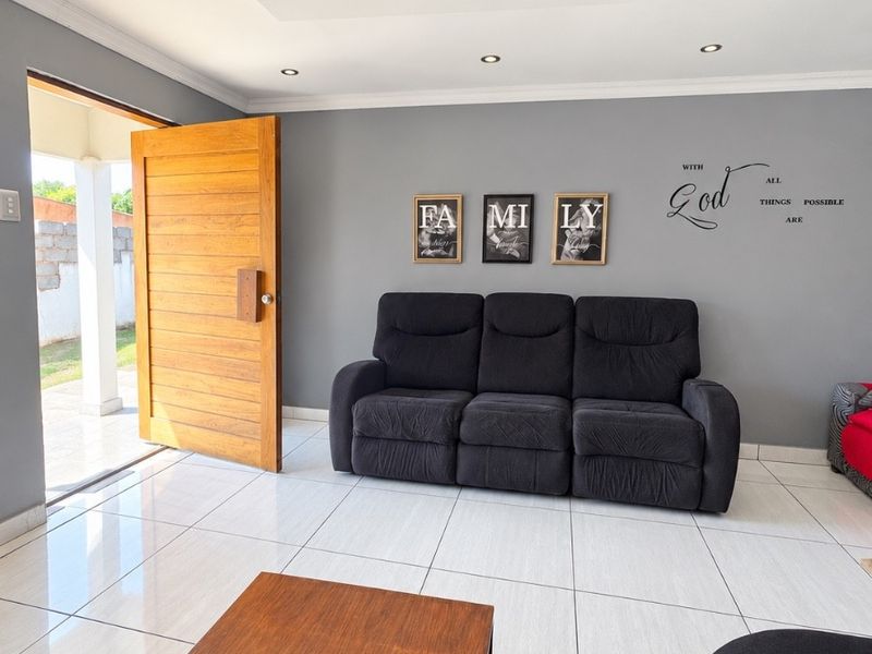 A bright living room featuring large format floor tiles, recessed ceiling lighting, and a solid wood entrance door leading to an outdoor patio area.