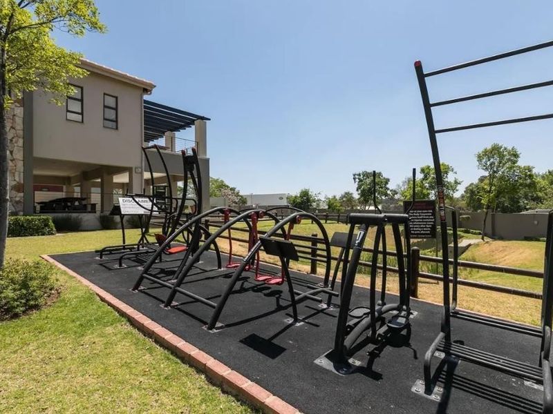 Outdoor gym facility featuring fixed exercise equipment on a rubberized surface, set within a well-maintained landscaped garden area.