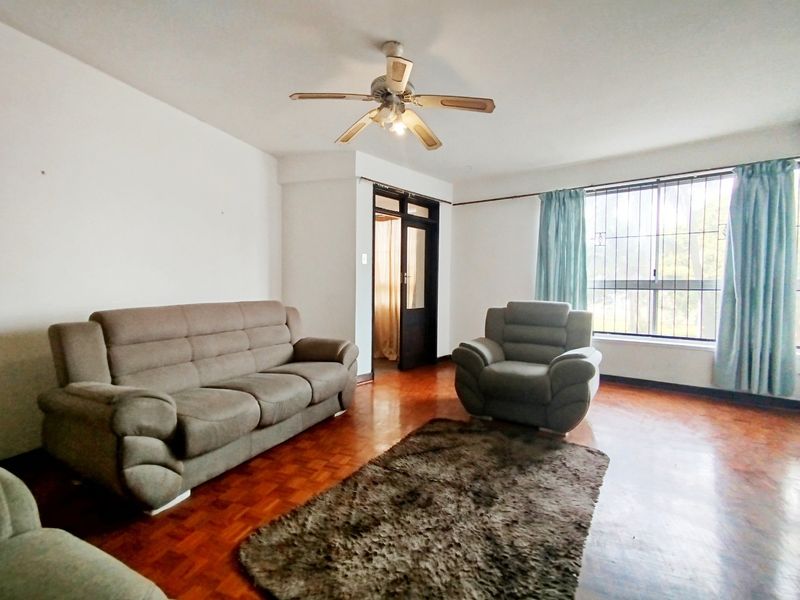A living room featuring parquet flooring, a ceiling fan, and large windows with security bars, offering a functional space with potential for modernization.