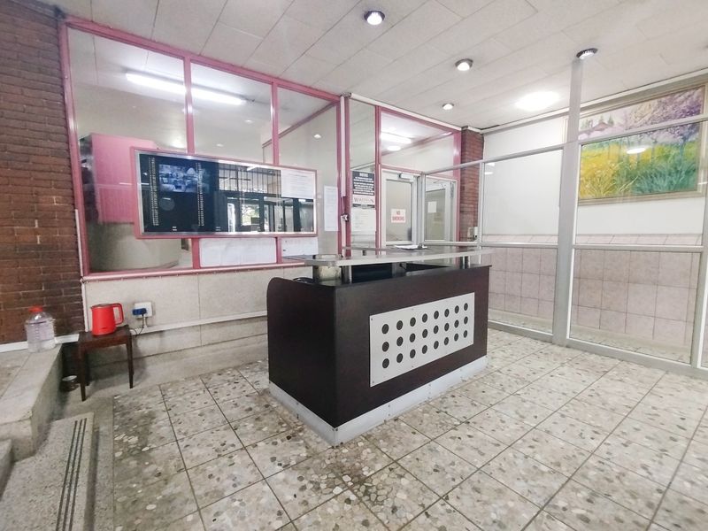 Building entrance lobby featuring a reception desk, glass partitions, and tiled flooring in a functional, dated commercial style.