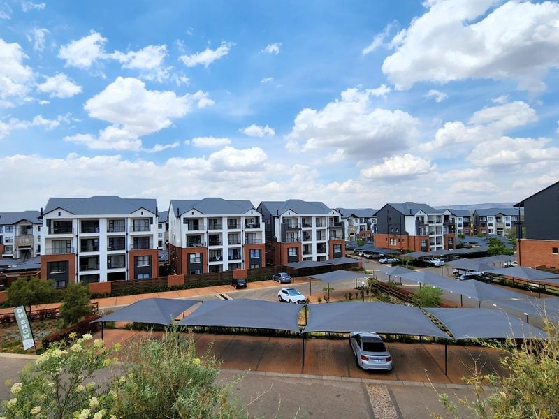 An elevated aerial view of a modern apartment complex featuring multiple residential blocks, paved parking areas, and shade cloth carports.