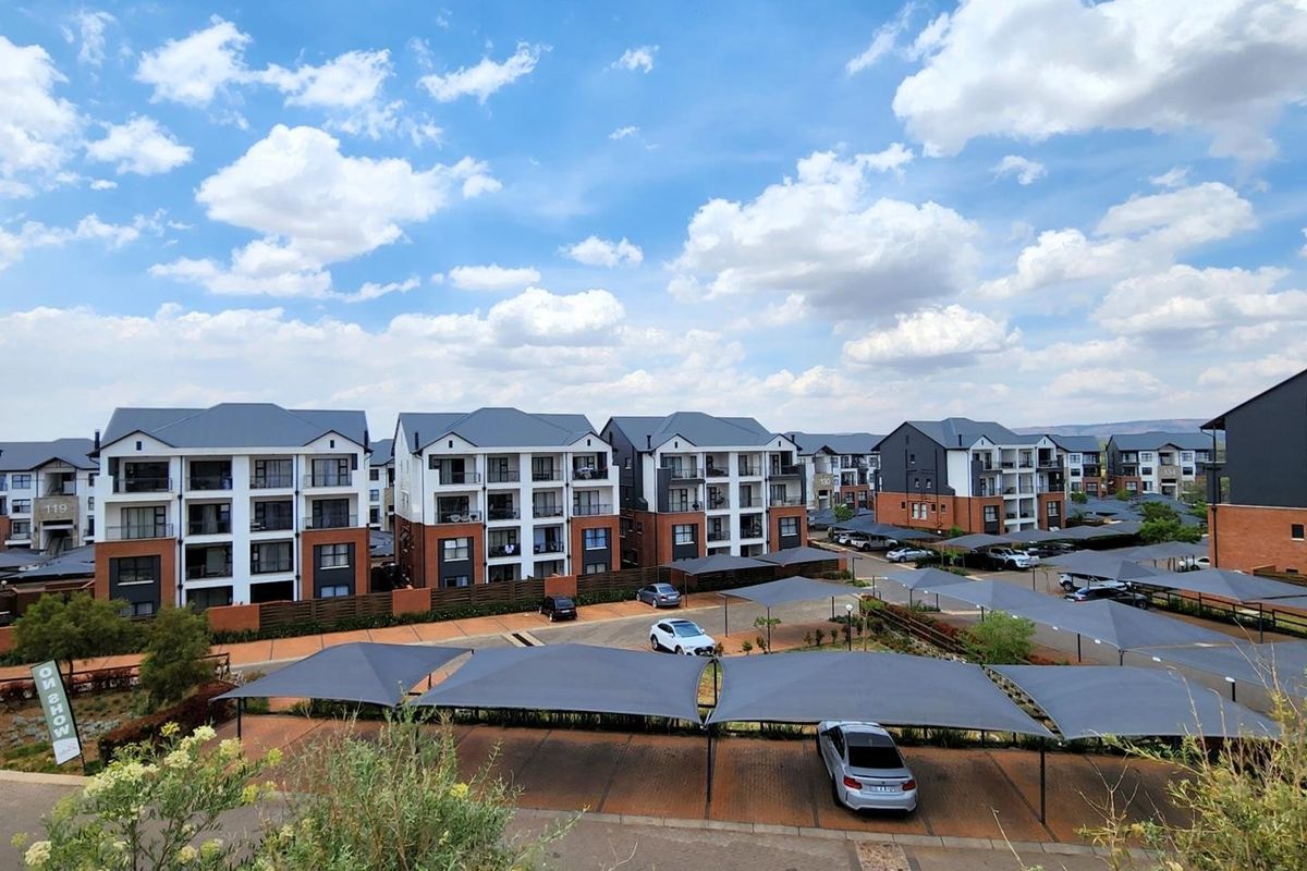 An elevated aerial view of a modern apartment complex featuring multiple residential blocks, paved parking areas, and shade cloth carports.