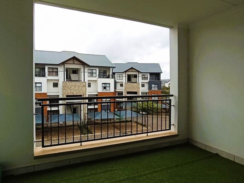 A covered balcony featuring a metal balustrade and artificial grass flooring, offering an open view of the apartment complex grounds.