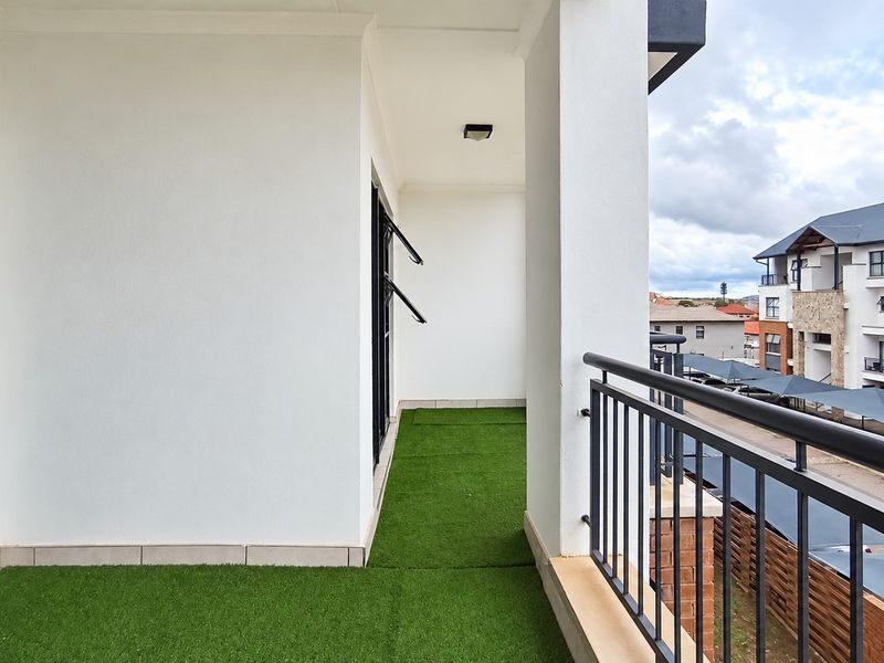 A clean, covered balcony featuring artificial grass flooring, a metal safety balustrade, and views over the apartment complex.