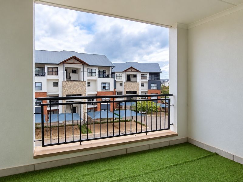 A clean, covered balcony featuring a metal balustrade and artificial grass flooring, offering an open view of the apartment complex grounds.