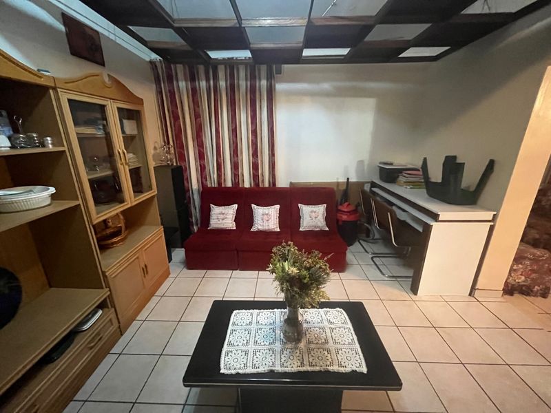 A living room featuring tiled flooring and a coffered ceiling, currently furnished with dated cabinetry and seating.
