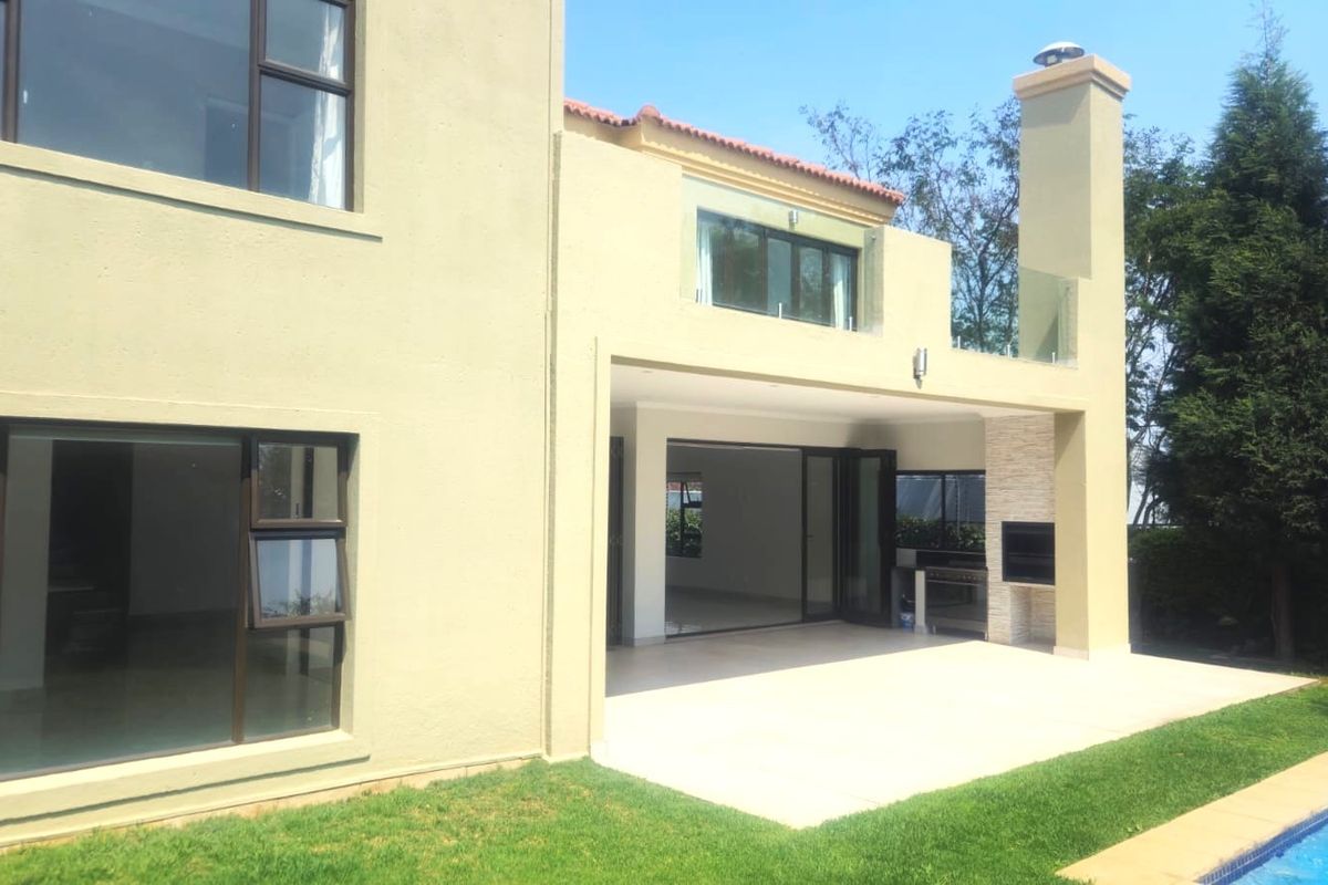 Rear elevation featuring a covered patio with a built-in braai, glass balustrade balcony above, and access to a swimming pool.