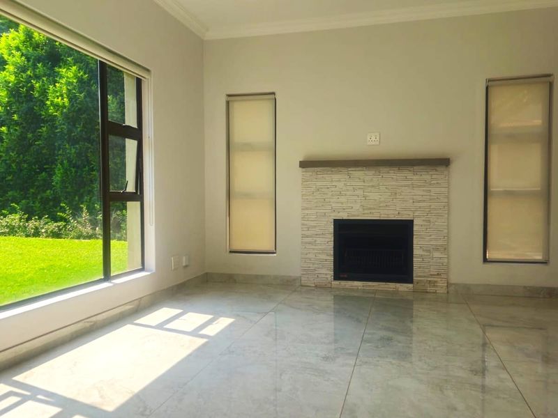 A bright, empty living room featuring polished tiled floors, a stone-clad fireplace, and large windows overlooking a lush garden.