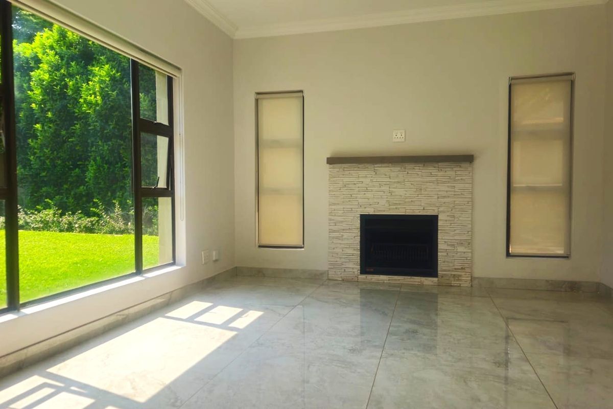 A bright, empty living room featuring polished tiled floors, a stone-clad fireplace, and large windows overlooking a lush garden.