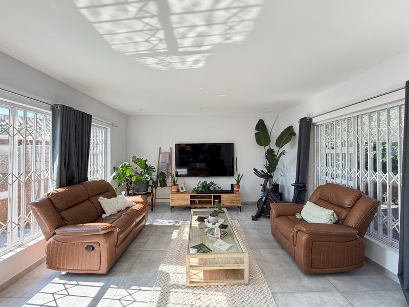 A bright and spacious living room featuring large windows with security gates, neutral tiled flooring, and recessed downlighting.