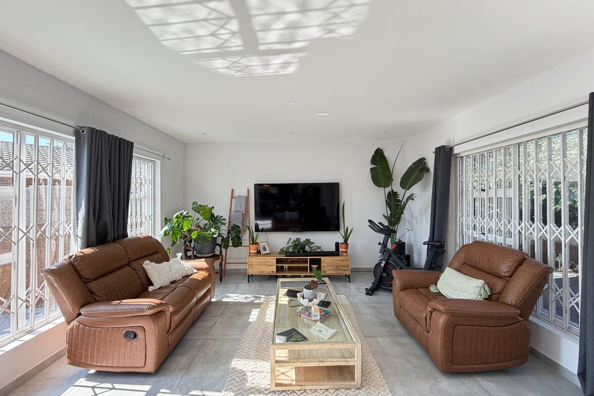 A bright and spacious living room featuring large windows with security gates, neutral tiled flooring, and recessed downlighting.