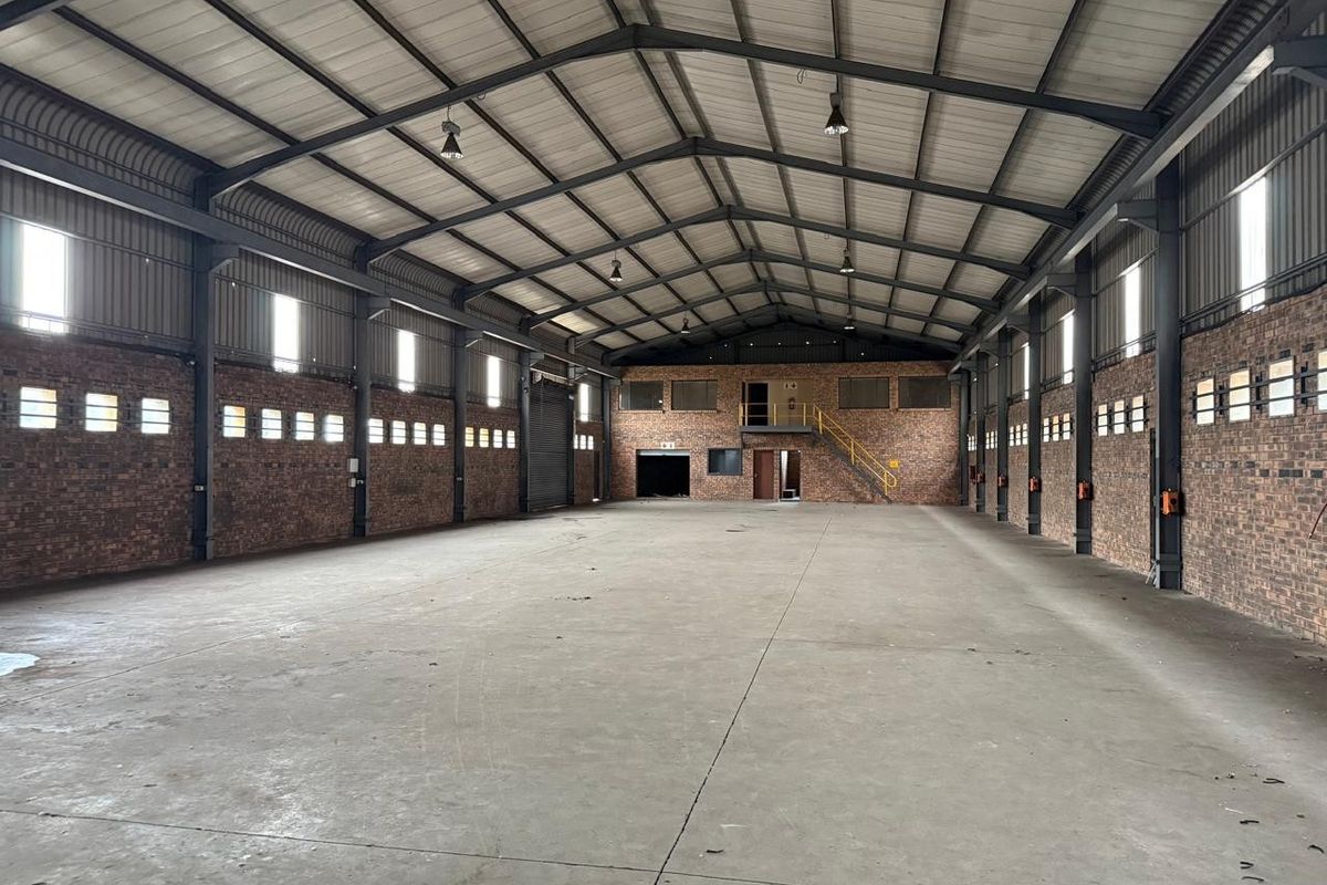 Spacious industrial warehouse featuring high ceilings with exposed steel trusses, concrete flooring, face brick walls, and a mezzanine level with staircase access.