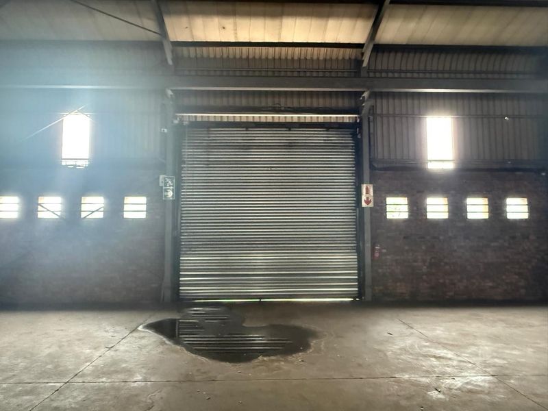 Spacious industrial warehouse or garage area featuring a large roller shutter door, exposed steel roof structure, and durable concrete flooring.