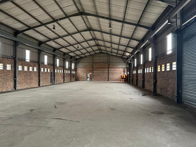 Spacious industrial warehouse featuring exposed brick walls, high-clearance steel portal frame, concrete flooring, and high-bay lighting, suitable for storage or manufacturing operations.