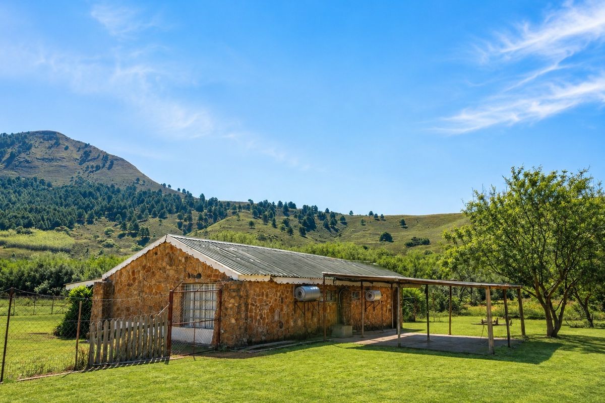 A rustic stone-clad farmhouse exterior featuring a corrugated metal roof, a covered patio area, and expansive surrounding green lawns with mountain views.